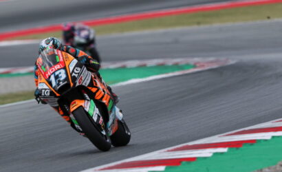 Racer in orange-and-black suit leans into a sharp turn on a race track, bike number 13 adorned with sponsor stickers.