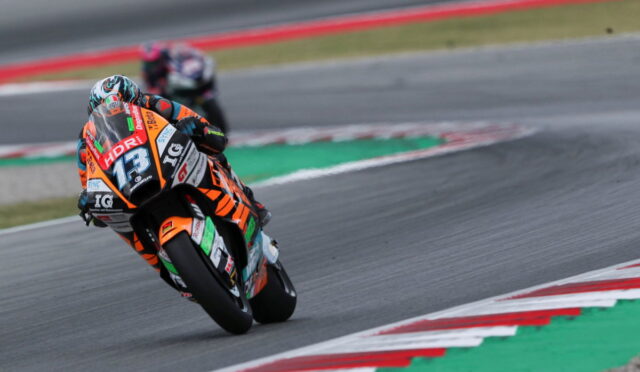 Racer in orange-and-black suit leans into a sharp turn on a race track, bike number 13 adorned with sponsor stickers.