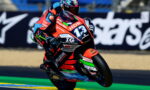 Motorcycle racer in red and black leathers leaning into a turn on a racing bike with number 13 and sponsor logos.