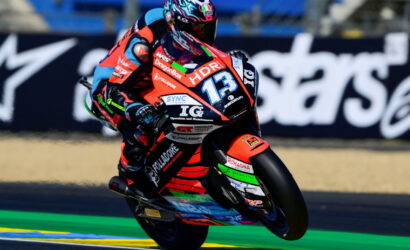 Motorcycle racer in red and black leathers leaning into a turn on a racing bike with number 13 and sponsor logos.
