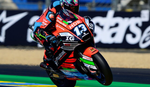 Motorcycle racer in red and black leathers leaning into a turn on a racing bike with number 13 and sponsor logos.