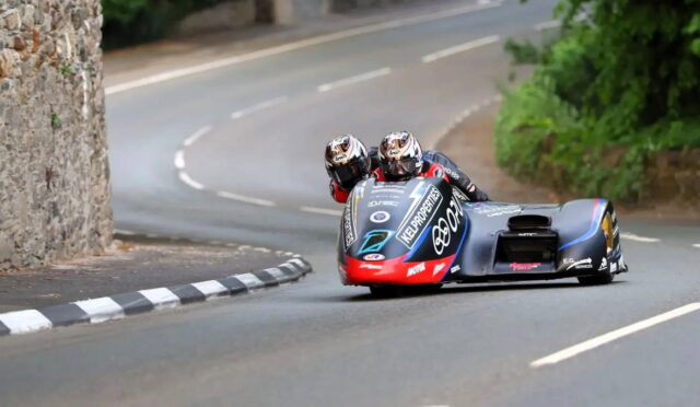Two riders on a sidecar motorcycle lean into a sharp turn on a winding road beside a stone wall.