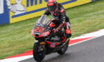 Racer in black-red suit leans into a corner on a red-and-black motorcycle labeled 22, speeding on a race track with yellow barriers in the background.