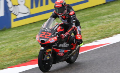 Racer in black-red suit leans into a corner on a red-and-black motorcycle labeled 22, speeding on a race track with yellow barriers in the background.