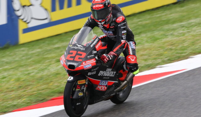 Racer in black-red suit leans into a corner on a red-and-black motorcycle labeled 22, speeding on a race track with yellow barriers in the background.