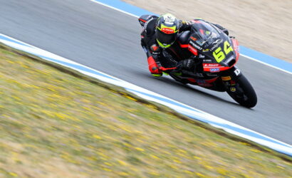 Motorcycle racer leaning into a sharp turn on a race track, number 64 bike in black and red livery ready for a cornering maneuver.