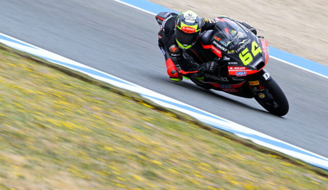 Motorcycle racer leaning into a sharp turn on a race track, number 64 bike in black and red livery ready for a cornering maneuver.
