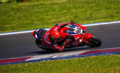 Red Honda motorcycle racer in a red helmet leans into a sharp corner on a race track.