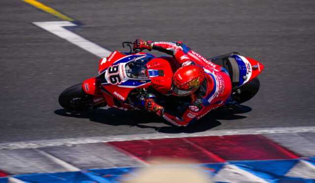 Motorcycle racer in red and blue livery leaning into a sharp turn on a race track.