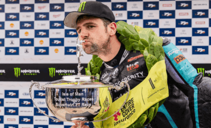 Man wearing a cap and laurel wreath kisses a large silver trophy, backdrop of sponsor logos visible (Isle of Man TT race)