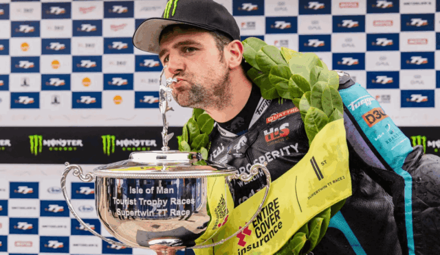 Man wearing a cap and laurel wreath kisses a large silver trophy, backdrop of sponsor logos visible (Isle of Man TT race)