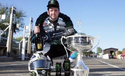 Racer kneels on a track posing with a large silver trophy, wearing a racing suit and Monster cap, holding a champagne bottle and a decorative wreath nearby.
