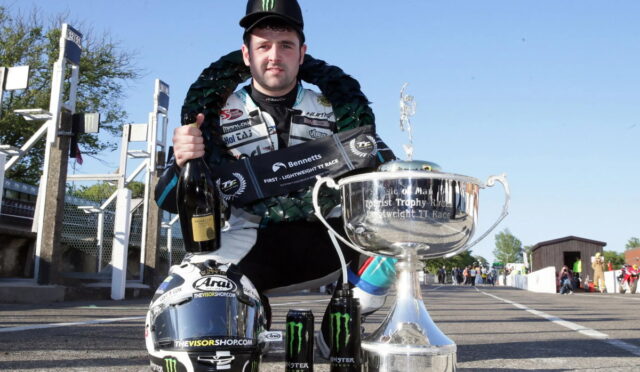 Racer kneels on a track posing with a large silver trophy, wearing a racing suit and Monster cap, holding a champagne bottle and a decorative wreath nearby.