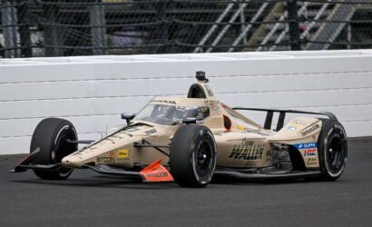 Open-wheel race car in beige livery rounds a track corner, large rear wing and exposed wheels, white barrier in background with chain-link fence above.
