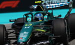 Green Formula 1 car speeding on a race track, driver in blue helmet with sponsor logos visible on the body.