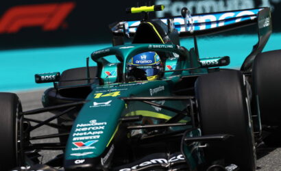 Green Formula 1 car speeding on a race track, driver in blue helmet with sponsor logos visible on the body.