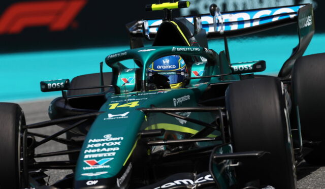 Green Formula 1 car speeding on a race track, driver in blue helmet with sponsor logos visible on the body.