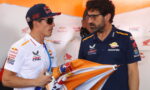 Two men in Honda Repsol racing gear chat in the pit area; one wears a Red Bull cap and sunglasses, holding a water bottle and a flag/cloth.
