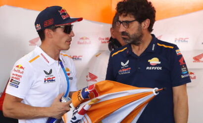 Two men in Honda Repsol racing gear chat in the pit area; one wears a Red Bull cap and sunglasses, holding a water bottle and a flag/cloth.