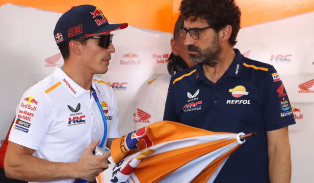 Two men in Honda Repsol racing gear chat in the pit area; one wears a Red Bull cap and sunglasses, holding a water bottle and a flag/cloth.