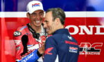 Rider in red Honda racing suit and white cap laughs while shaking hands with a teammate in a blue shirt against a bold red and blue Honda backdrop.