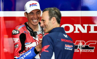 Rider in red Honda racing suit and white cap laughs while shaking hands with a teammate in a blue shirt against a bold red and blue Honda backdrop.