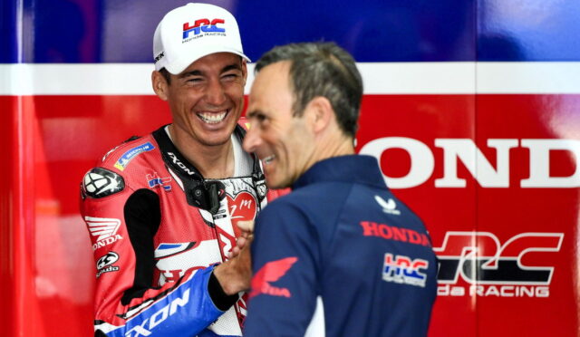 Rider in red Honda racing suit and white cap laughs while shaking hands with a teammate in a blue shirt against a bold red and blue Honda backdrop.