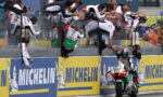 Rider on a green, white and red motorcycle is greeted by a cheering crowd leaning over the barrier at the track's edge, Michelin ads in view.