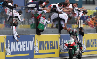 Rider on a green, white and red motorcycle is greeted by a cheering crowd leaning over the barrier at the track's edge, Michelin ads in view.