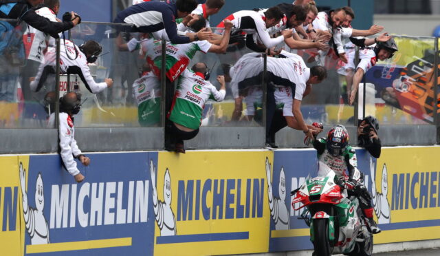 Rider on a green, white and red motorcycle is greeted by a cheering crowd leaning over the barrier at the track's edge, Michelin ads in view.