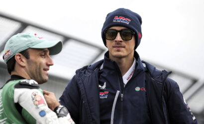 Two race car drivers converse outdoors; one in a light cap and racing suit, the other in a navy jacket, beanie and sunglasses.
