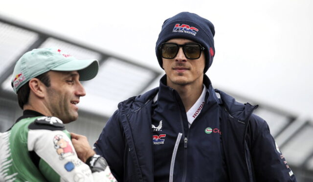 Two race car drivers converse outdoors; one in a light cap and racing suit, the other in a navy jacket, beanie and sunglasses.