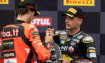 Two MotoGP riders in team racing suits shake hands on a podium with Pirelli banners in the background.