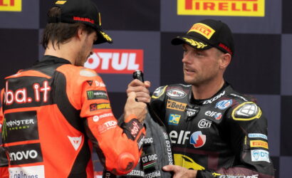 Two MotoGP riders in team racing suits shake hands on a podium with Pirelli banners in the background.