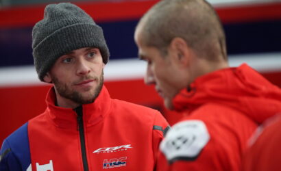 Two men in red racing jackets talk closely, one wearing a gray knit beanie and looking at the other man.