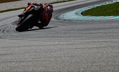 Motorcycle racer in orange gear leans into a left-hand turn on a race track, blue-striped run-off and grassy edge in the background.