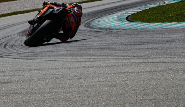 Motorcycle racer in orange gear leans into a left-hand turn on a race track, blue-striped run-off and grassy edge in the background.