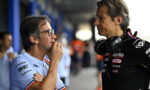 Two men in a motorsport paddock talk; one wears a light-blue polo with glasses, the other a black Aprilia racing suit with headphones around his neck.