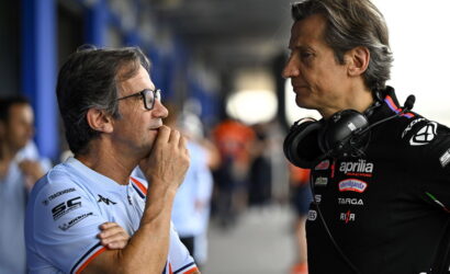 Two men in a motorsport paddock talk; one wears a light-blue polo with glasses, the other a black Aprilia racing suit with headphones around his neck.