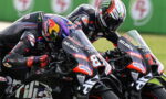 Two professional motorcycle racers lean into a tight turn on parallel racing bikes, helmets and black-red leathers screaming speed on a track backdrop.