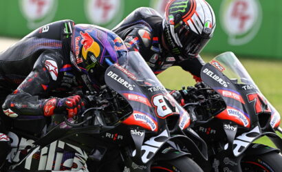 Two professional motorcycle racers lean into a tight turn on parallel racing bikes, helmets and black-red leathers screaming speed on a track backdrop.