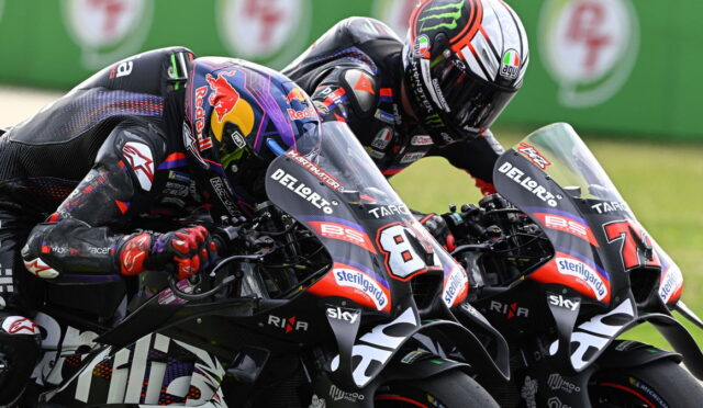 Two professional motorcycle racers lean into a tight turn on parallel racing bikes, helmets and black-red leathers screaming speed on a track backdrop.