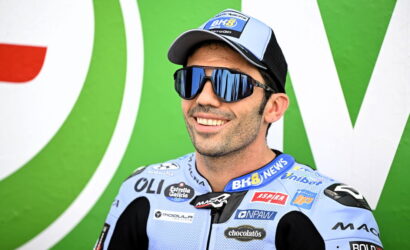 Male motorcycle racer smiling in a light blue suit with sponsor logos, blue mirrored sunglasses, and a cap, against a green backdrop.