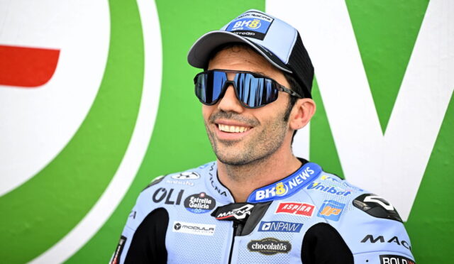 Male motorcycle racer smiling in a light blue suit with sponsor logos, blue mirrored sunglasses, and a cap, against a green backdrop.
