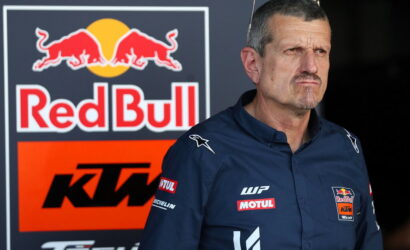 Man in a blue team shirt with Red Bull and KTM logos standing near a Red Bull KTM backdrop in a paddock area.