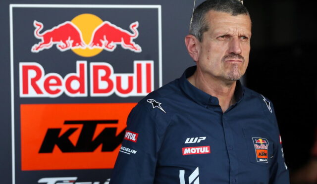 Man in a blue team shirt with Red Bull and KTM logos standing near a Red Bull KTM backdrop in a paddock area.