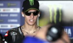 Moto racer wearing a black Monster Energy cap and dark sunglasses speaks to the camera during a media interview, wearing earphones and a star-studded necklace over a Yamaha racing suit.