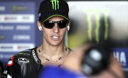 Moto racer wearing a black Monster Energy cap and dark sunglasses speaks to the camera during a media interview, wearing earphones and a star-studded necklace over a Yamaha racing suit.