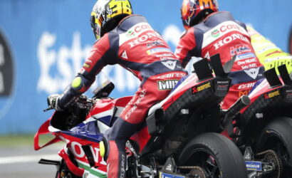 Two motorcycle racers on red Honda bikes leaning into a turn with a blue barrier behind them.