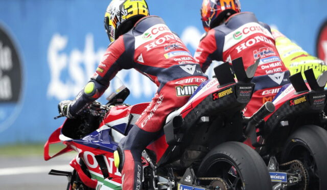 Two motorcycle racers on red Honda bikes leaning into a turn with a blue barrier behind them.